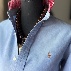 May include: A light blue button-down shirt featuring a red and white gingham pattern on the inner collar. The shirt displays a small embroidered logo of a horse and rider on the left chest and is styled with a dark brown beaded necklace.