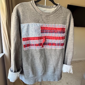 May include: A gray sweatshirt with a patchwork American flag design on the front. The flag features a blue floral print for the stars and red and white plaid stripes. The sleeves are cuffed, revealing a lighter fabric.