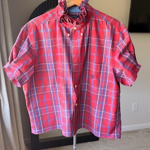 Op de afbeelding: Rood en blauw geruit shirt met korte mouwen en een gerimpelde kraag. Het shirt heeft een knoopsluiting aan de voorkant en een klein logo op de borst. Het shirt hangt aan een houten hanger.