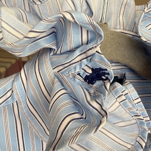 Könnte beinhalten: Nahaufnahme eines hellblau-weiß gestreiften Hemdes mit einem marineblauen, gestickten Logo. Das Hemd hat einen Rüschenkragen und hängt an einem grauen Kleiderbügel.