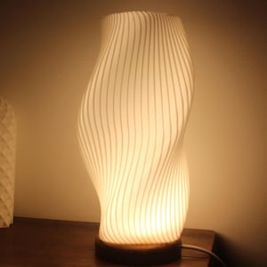 Può includere: Una lampada bianca a strisce verticali con una forma curva a clessidra. La lampada è illuminata, emettendo un bagliore caldo. Poggia su una base di legno scuro ed è collegata. Un secondo oggetto geometrico è parzialmente visibile a sinistra.