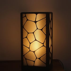 Peut inclure: Lampe de table rectangulaire avec un cadre sombre et un motif. L'intérieur de la lampe brille d'une lumière jaune chaude, créant une ambiance chaleureuse. Le design présente un réseau de formes irrégulières.