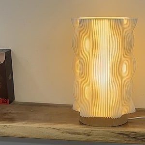 Peut inclure: Une lampe de table blanche à motifs ondulés dégageant une lumière chaude. La lampe a une forme cylindrique avec des lignes verticales et repose sur une base ronde de couleur claire. La lampe est branchée.