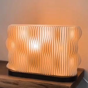 Peut inclure: Une lampe de table crème moderne avec un design unique de lignes verticales ondulées. La lampe a une forme rectangulaire avec des bords arrondis et une base noire. La lumière brille chaleureusement de l'intérieur, créant une ambiance douce.