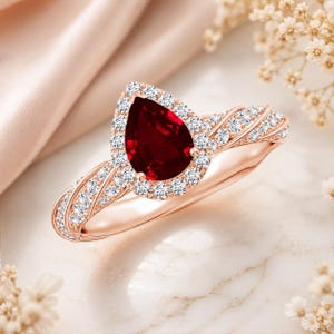 Pode incluir: Anel de ouro rosa com uma pedra preciosa vermelha escura em forma de pêra, rodeada por pequenas pedras preciosas transparentes. A banda também é adornada com pedras preciosas transparentes, criando um efeito brilhante. O anel é apresentado sobre um fundo suave e neutro.