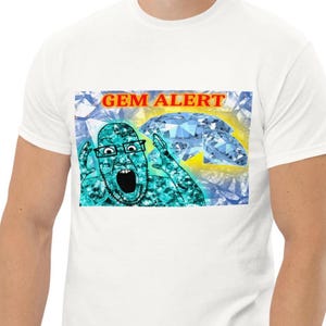 以下が含まれることがあります： 白いTシャツで、口を開けた表情で眼鏡をかけ、青いダイヤモンドの模様で覆われた人物のグラフィックプリントが施されています。人物の上には赤い文字で「GEM ALERT」と書かれており、背景には大きな青いダイヤモンドが描かれています。