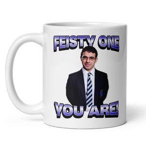 May include: White ceramic mug featuring a man in a suit and tie with the text "FEISTY ONE YOU ARE!" in blue and black lettering. The mug has a curved handle and a glossy finish.