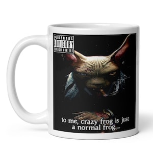 May include: White ceramic mug featuring a surreal illustration of a frog-like creature smoking a cigar. The mug includes the text "to me, crazy frog is just a normal frog..." and a "Parental Advisory Explicit Content" label.