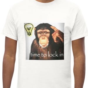 Peut inclure: Un t-shirt blanc à manches courtes avec une impression graphique représentant un chimpanzé pointant sa tête avec une icône d'ampoule jaune au-dessus. Le texte "time to lock in" est imprimé en lettres blanches au bas du motif carré.