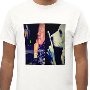 Puede incluir: Una camiseta blanca de manga corta con un estampado gráfico cuadrado en el pecho. El gráfico está dividido en dos paneles verticales, mostrando a una persona a la izquierda y a un perro a la derecha, ambos haciendo un gesto de silencio.