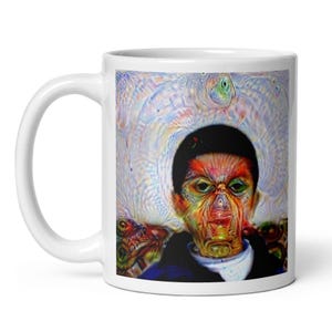 May include: A white ceramic mug featuring a colourful, abstract, and surreal graphic print. The design displays a central figure with a dark, rounded head, textured facial features in shades of orange, green, and yellow, and a swirling, light-coloured halo.