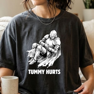 Może przedstawiać: Ciemnoszary t-shirt z krótkim rękawem z białą grafiką przedstawiającą rycerza w pełnej zbroi, siedzącego z dłońmi na brzuchu. Poniżej ilustracji rycerza znajduje się napis "TUMMY HURTS" wykonany białymi wielkimi literami.