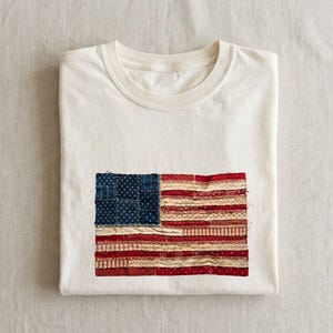 Puede incluir: Camiseta crema doblada con un diseño de bandera estadounidense cosido. La bandera presenta un cuadrado azul con estrellas blancas y rayas rojas y blancas. La camiseta está cuidadosamente doblada, mostrando los detalles de la bandera.