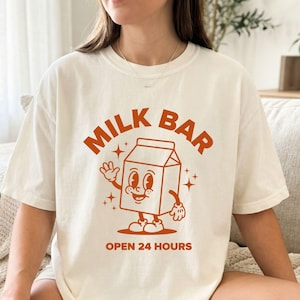 Peut inclure: Un t-shirt à manches courtes blanc cassé avec une impression graphique orange. Le motif représente un personnage de brique de lait avec des bras et des jambes, entouré d'étoiles. Le texte indique MILK BAR en arc et OPEN 24 HOURS en dessous.
