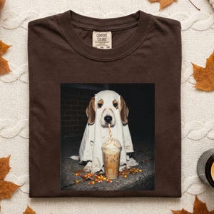 May include: A dark brown t-shirt featuring a graphic print of a dog wearing a white ghost costume, drinking an iced coffee with a striped straw. The dog is sitting on a dark surface with scattered candy corn. The shirt has a Comfort Colors label.