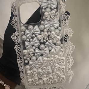 Può includere: Custodia per telefono trasparente decorata con fiocchi e perle bianche. La custodia ha un bordo in pizzo bianco e il testo "@casemyjunk". I fiocchi e le perle sono disposti sul retro della custodia.