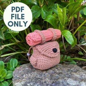 May include: A crocheted miniature backpack in shades of pink and brown, with a rolled-up sleeping bag attached. The backpack has a flap and a black button closure. The text "PDF FILE ONLY" is in a white circle in the background.