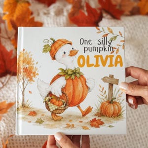 May include: A square children's book with a white cover featuring a watercolor illustration of a white duck wearing a pumpkin costume and hat. The duck carries a small basket with a pumpkin. Text reads One silly pumpkin OLIVIA near a wooden sign.