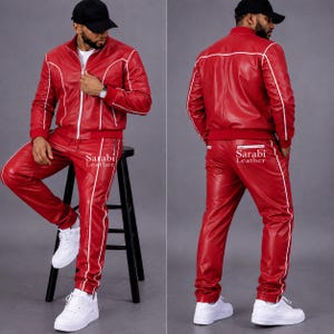 May include: A red leather tracksuit featuring a zip-up jacket and matching pants with white piping accents. The jacket has a stand-up collar and long sleeves. The pants have a straight-leg fit. The text Sarabi Leather is printed on the pants.