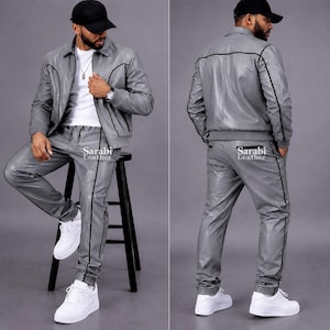 May include: A grey leather jacket and matching trousers set featuring black piping details. The jacket has a collar, long sleeves, and a front zipper. The trousers have a straight-leg fit. The text Sarabi Leather is printed on the clothing.