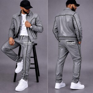 May include: A grey leather jacket and matching trousers set featuring black piping details. The jacket has a collar, long sleeves, and a front zipper. The trousers have a straight-leg fit. The text Sarabi Leather is printed on the clothing.