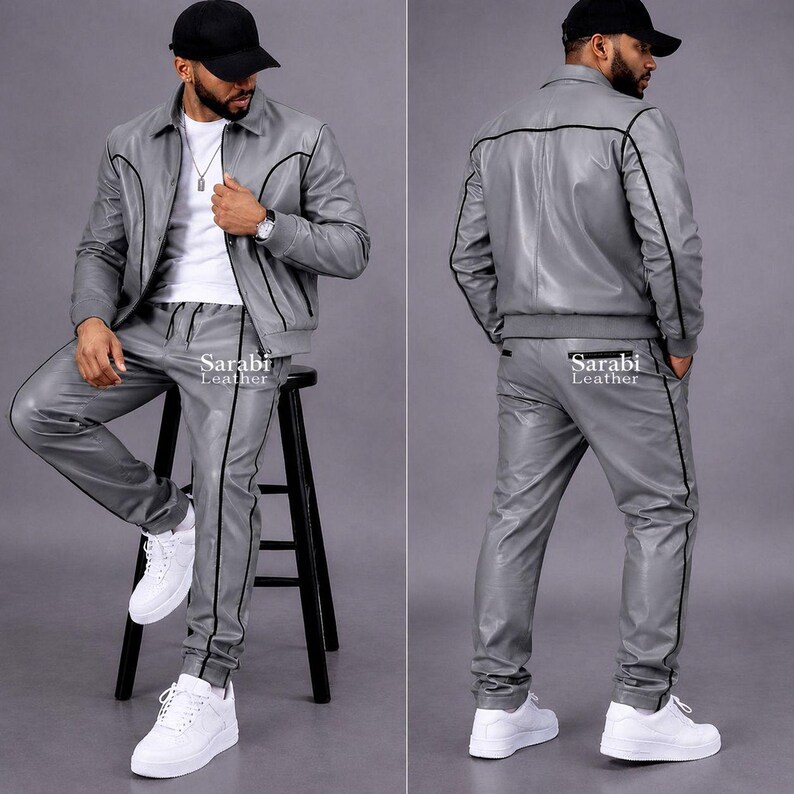 May include: A grey leather jacket and matching trousers set featuring black piping details. The jacket has a collar, long sleeves, and a front zipper. The trousers have a straight-leg fit. The text Sarabi Leather is printed on the clothing.