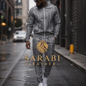 May include: A gray leather tracksuit with white piping, consisting of a zip-up bomber jacket and matching pants. The pants feature a gold lion logo and the text "SARABI LEATHER". The outfit is paired with white sneakers.