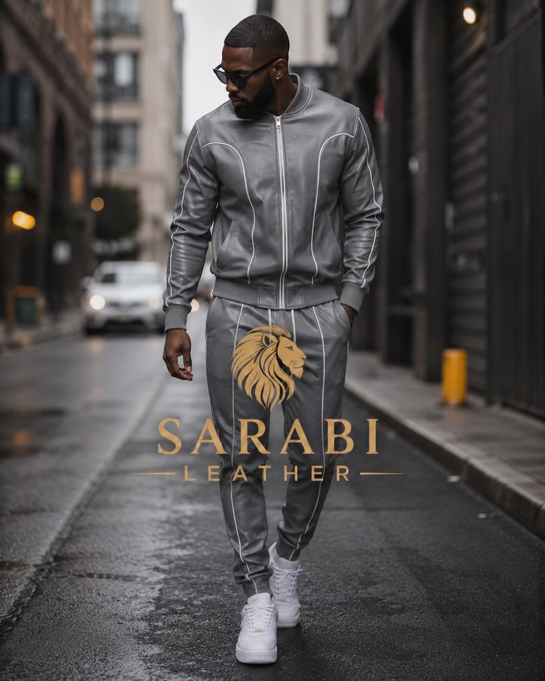 May include: A gray leather tracksuit with white piping, consisting of a zip-up bomber jacket and matching pants. The pants feature a gold lion logo and the text "SARABI LEATHER". The outfit is paired with white sneakers.