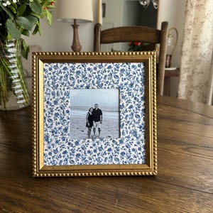 May include: A square gold-toned picture frame with a blue floral patterned mat. The frame holds a black and white photograph of two people. The frame sits on a wooden table, with a vase of flowers in the background.