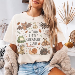May include: A cream-colored short-sleeve t-shirt featuring a graphic print of various small animals including a bat, frog, snail, and anglerfish, with the text "WEIRD LITTLE CREATURE CLUB" centered in black lettering among small leaf and star accents.