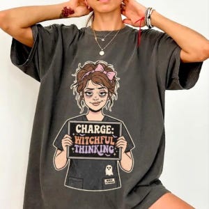 May include: A charcoal gray t-shirt featuring a graphic of a person with messy brown hair and a pink bow, holding a sign that reads "CHARGE: WITCHFUL THINKING" with a small ghost icon below. The design includes stars and bats in a retro-style font.