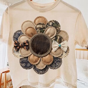 May include: A cream-colored short-sleeve t-shirt featuring a large, circular flower-shaped applique on the chest. The flower petals are made of various fabrics including tan suede, camouflage, burlap, and black paisley, with a dark brown fur center.