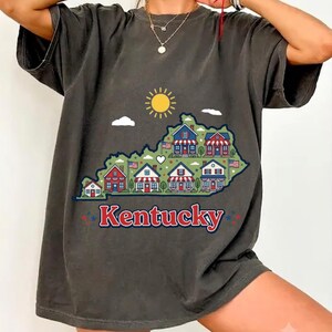 May include: A charcoal gray short-sleeve t-shirt featuring a graphic of the state of Kentucky outlined in green. Inside the outline are several houses with red, white, and blue patriotic decor, a yellow sun, and the word Kentucky in red text.