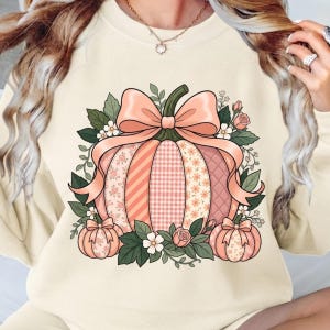 May include: A cream-colored crewneck sweatshirt featuring a large central graphic of a pumpkin decorated with various pink and peach patterns, including stripes, gingham, and florals, topped with a large bow and surrounded by green leaves and flowers.