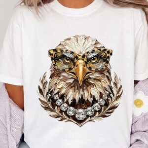 May include: A white t-shirt featuring a graphic of an eagle head with a textured, painterly appearance. The eagle wears leopard-print glasses and a necklace made of large, clear, faceted crystals. The design uses shades of brown, gold, and white.