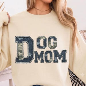 May include: A cream-coloured crewneck sweatshirt featuring the text DOG MOM in large block letters. The letters are constructed with a dark denim-like fabric border, a frayed edge, and a green and brown camouflage pattern fill inside each letter.