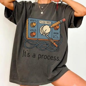 Pode incluir: Uma t-shirt cinzento-carvão de manga curta com um gráfico de um painel de controlo de sintetizador azul com botões cor de laranja, um lápis e um ferro de soldar. O texto "It's a process." está impresso a preto sob a ilustração do dispositivo.