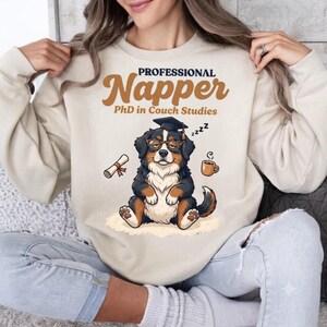 Pode incluir: Uma camisola bege de gola redonda com o gráfico de um cão de montanha de Berna a usar um chapéu de finalista e óculos. O texto diz Professional Napper PhD in Couch Studies, acompanhado por ilustrações de um diploma e uma caneca.