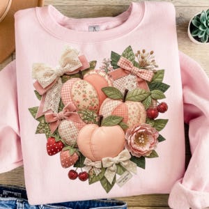 May include: A light pink sweatshirt featuring a central graphic of peaches, strawberries, and a flower, accented with fabric bows, lace, and green leaves. The design incorporates various patterns including gingham and floral prints in soft peach and red tones.