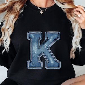 Puede incluir: Una sudadera negra de manga larga con una letra K grande centrada. La letra tiene una textura de tela vaquera azul con detalles desgastados y un borde blanco cosido, imitando la apariencia de un parche de mezclilla aplicado.