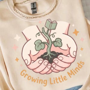 Op de afbeelding: Een beige sweater met ronde hals en een afbeelding van twee komvormige handen die een plant vasthouden met een boek als bovenste blad. De tekst "Growing Little Minds" is in een oranje boog onder de handen gedrukt, omringd door sterretjes.