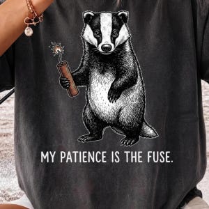 May include: A charcoal grey t-shirt featuring a black and white illustration of a badger holding a lit stick of dynamite. Below the badger, the text reads, "MY PATIENCE IS THE FUSE." The design is centred on the front of the garment.