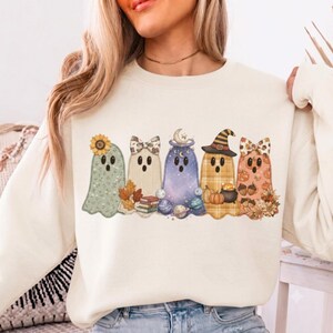 May include: A cream-colored crewneck sweatshirt featuring a graphic print of five cartoon ghosts. The ghosts are decorated with various patterns and items including a sunflower, a bow, books, planets, a witch hat, pumpkins, and acorns.