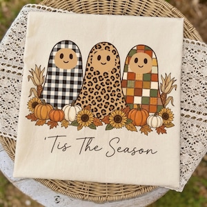 May include: A cream-coloured t-shirt featuring a graphic of three ghosts with smiling faces, wearing patterns of black and white buffalo plaid, leopard print, and autumn-coloured patchwork. Below are pumpkins, sunflowers, and the text 'Tis The Season'.