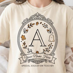 May include: A cream-coloured t-shirt featuring a black circular frame design with a large letter A in the centre. The design includes a puzzle piece, a ghost, and acorns, with a banner reading Your Name Here and the text SPECIAL EDUCATION TEACHER.