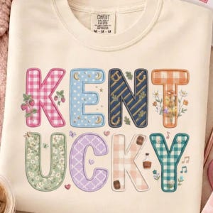 Puede incluir: Una camiseta de color crema con la palabra KENTUCKY en letras grandes y estampadas. Cada letra muestra diseños únicos, incluyendo cuadros vichy rosas con fresas, lunares azules, rayas azul marino con herraduras y estampados florales.