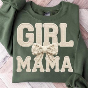 May include: An olive green crewneck sweatshirt featuring the text GIRL MAMA in large, cream-colored, textured varsity-style letters. A cream-colored, three-dimensional knit bow is positioned directly between the two words on the front.