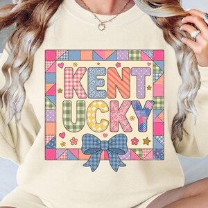 Puede incluir: Una sudadera de color crema que presenta un gráfico central con el texto KENTUCKY en letras de bloque. Las letras y el borde circundante están rellenos con varios patrones coloridos de estilo patchwork, incluyendo cuadros, flores y lunares, con un lazo azul.