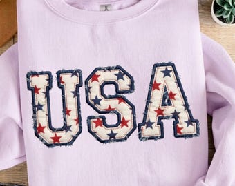 May include: A light purple crewneck sweatshirt featuring the letters USA in a large, block-style font. The letters are filled with a cream-colored fabric patterned with small red and navy blue stars, finished with frayed, dark blue stitched edges.