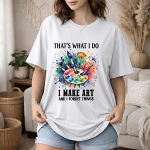 Könnte beinhalten: Ein weißes Kurzarm-T-Shirt mit einer bunten Grafik einer Künstlerpalette und verschiedenen Pinseln, umgeben von Aquarellspritzern. Der Text lautet: THAT'S WHAT I DO I MAKE ART AND I FORGET THINGS in schwarzen Großbuchstaben.